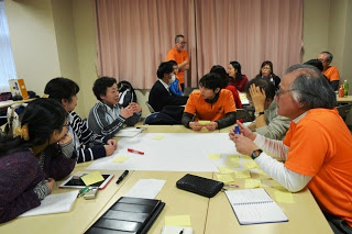 オレンジ色のシャツを着たファシリテーターが参加者のサポートをし、男女・年齢問わず多様な人々が参加しながらグループディスカッションをしている様子を写した写真