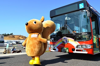 黄色い長靴を履いたリスのキャラクターの着ぐるみが、虹が描かれた赤色のJRバスの前に立ちポーズをとっている写真