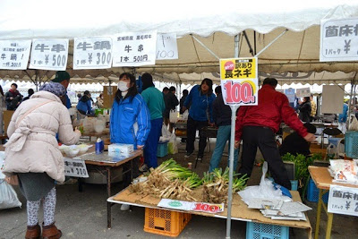 直売所のテントの下で根っこがついた長ねぎが100円で販売されており、左側の牛鍋のブースで来場者が店員とやりとりしている様子の写真