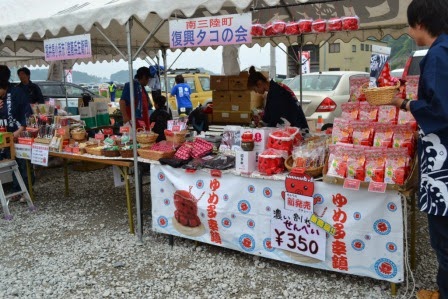 「南三陸町復興タコの会」と書かれたテント下の屋台に、オクトパス君のイラストが描かれた沢山のお菓子や土産品が並び、紺色の法被姿の販売員が商品を並べている様子の写真