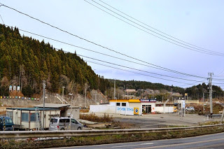 森を背景に、黄色い屋根のコイン洗車場やプレハブ小屋が道路沿いに並んでいる様子の写真