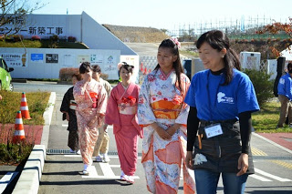 青いTシャツの案内係が先導している通路を、色とりどりの着物を着た女性たちが笑顔で歩いている様子の写真
