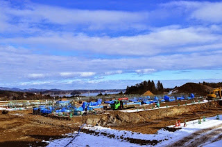 雪が少し残り、中央の盛られた土の上の所々がブルーシートで囲われた建設現場の写真