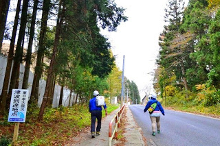 ヘルメットをかぶった2人の男性が「津波避難地点」の看板がある坂道の歩道を歩いて避難訓練に参加している様子の写真