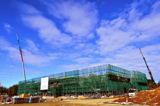 青空の下、広い建設現場で大型の建物が緑色の足場に覆われて建設中であり、左右に高いクレーン車が立ち、周囲には重機や建設資材が置かれている建設中の建物の写真