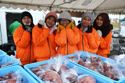 お揃いのオレンジ色の防寒ジャンパーを着た5人の女性たちが、笑顔で手を頬に当ててポーズを取り、手前には袋詰めされた赤いタコが並んだコンテナが置かれている鮮魚販売ブースの写真