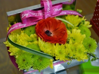 銀色に光沢のある長方形の器に、中央の赤い花がひときわ目を引き、そのまわりを黄色や黄緑の小菊のような花々と細長い緑の葉で美しくまとめられ、全体に濃いピンクのリボンがかけられた華やかなフラワーアレンジメントの写真