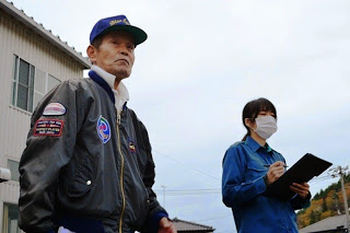 年配の男性が真剣な表情で前を見つめて立っており、その横で女性スタッフがマスクをつけてメモを取っている写真