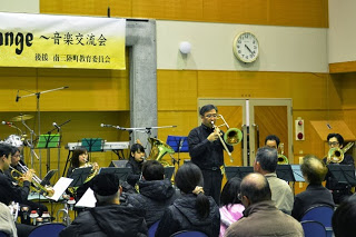 吹奏楽団の中年男性がトロンボーンでソロ演奏を行い、観客が座って静かに聴いている音楽交流会の様子の写真