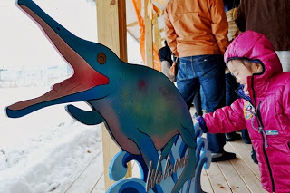 雪景色の中、ピンクの防寒着を着た子どもが、カラフルな魚竜の木製看板に触れて楽しそうにしている屋外の様子の写真