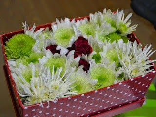 赤地に白い水玉模様の四角い箱のふたが外され、白や黄緑の花々の中に赤い小さなバラが3輪ほどアクセントとしてあしらわれ、花がぎっしりと詰め込まれた華やかなフラワーギフトボックスの写真