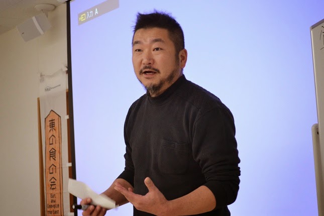 黒い服を着た男性がスクリーンの前に立ち、手に紙を持ちながら熱心に話をしている写真