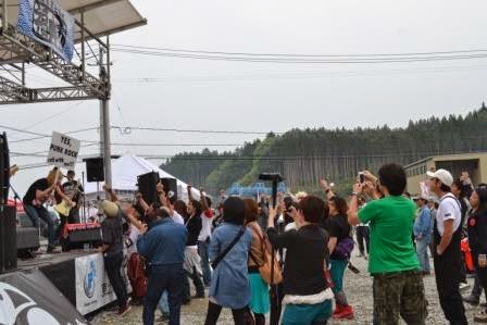 バンド演奏が行われたステージで、文字が書かれた白い看板を持つ人の前に観客たちが歓声を上げたり写真を撮ったりして盛り上がっている様子の写真