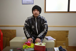 リボンがついた花束と赤い箱に入ったフラワーアレンジメンなどが置かれたテーブルの前に座っている黒いニット帽と黒のスポーツジャケットを着た小野政道さんの写真