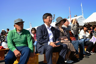 晴れた空の下、イベント用の簡易ベンチに座ったスーツ姿の男性や帽子をかぶった高齢者などが観客としてステージを見ている写真