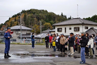 屋外の広場で青い制服姿の消防隊員の男性が拡声器を使って住民に向かって説明を行い、住民たちが真剣に聞いている様子の写真