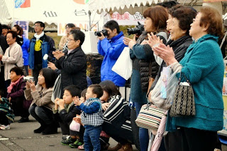 屋外のテント前で子どもから高齢者まで多くの観客が笑顔で拍手を送り、パフォーマンスを見守っている様子の写真