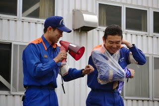 防災訓練中に拡声器を持った職員が、もう一人の職員の右腕に応急処置用のビニールをかける実演をしている写真