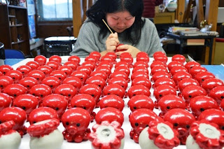 一人の女性が作業台の上に沢山並べられた赤いタコの置物に、丁寧に顔を描いている様子の写真