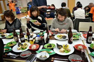 テーブルいっぱいに並べられた豪華な和食料理と瓶ビールやお茶、醤油などの調味料が並べられ参加者の女性たちが食事を楽しんでいる写真