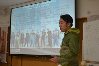 「FISHERMEN, BE AMBITIOUS!」と書かれたスライドの前で、緑の服を着た男性が漁業者に向けて話をしている写真