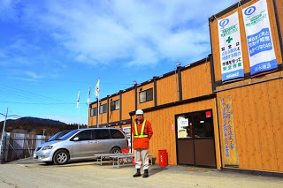 白いヘルメットをかぶりオレンジの作業着を着た男性が工事現場の詰所の前に立っている写真