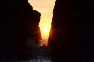 岩の間から昇ってきた朝日が中央に輝き、海面にオレンジ色の光を映し出しながら岩の輪郭をシルエットに浮かび上がらせる幻想的な海岸の写真