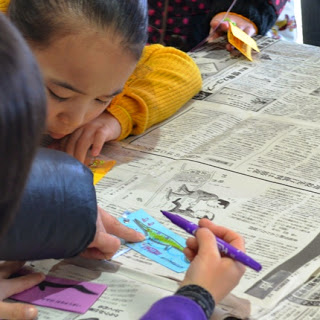 新聞紙が広げられたテーブルの上で子どもが、長方形の形の画用紙にペンで絵を描いている様子を写した写真