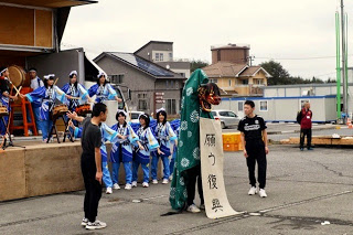 左右に黒いシャツを着た2人の男子生徒が立ち、中央の緑色の獅子舞が「願う復興」と書かれた垂れ幕をくわえ、お揃いの衣装を着た生徒たちが叩く太鼓の演奏に合わせてポーズを決めている様子の写真