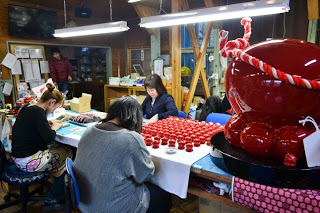 室内の作業場で数人の女性が赤いタコの置物を机いっぱいに並べて手作業で絵付けしている様子の写真