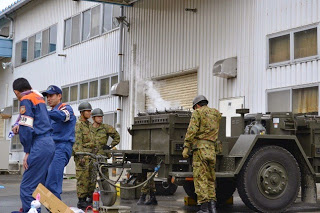 応急屋外で応急処置の実演を行っている2人の消防隊員の後ろで、迷彩柄の作業服を着た自衛隊員が、湯気が上がった大型の炊飯用トレーラーの前で準備をしている様子の写真