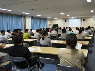 前方のスクリーンを使いながら講演をしている亀山さんと、会場の会議室にずらりと並んだ長机の席に多数の出席者が座っている様子を後方から写した写真