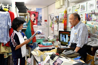 商品がたくさん並ぶ店舗内で、生徒が紙を手にして話しながら、カウンターの中にいる男性が笑顔で応えている様子が写っている写真