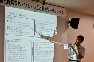 会議室で、男性が前方のスクリーンに映し出されたスライドを指し示しながら説明している様子の写真