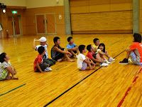 体育館の床に座って先生の話を聞いている子供たちの写真