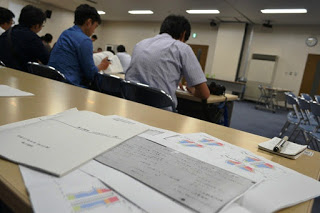 長机に向かって座った参加者たちの後ろ姿を、長机の上に複数の資料が広げられている様子越しに写した写真