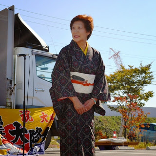 屋外ステージの前で、白い帯に濃い色の着物を着た年配の女性が、両手を前に重ねて笑顔で立っている写真