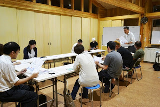 木の内装の部屋でホワイトボードの前に立つ男性の話を、コの字型に設置された長机を囲んだ参加者が資料を見ながら聞いている写真