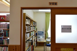 中央に「学習室」と書かれたプレート付きのドアがあり、そのガラス部分には「東日本大震災資料」と書かれた紙が貼られ、ドアの奥には本棚や資料、緑色のカーテンと机が見え、左側にも本棚が並び多くの本が置かれている写真