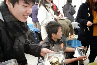 笑顔の若い男性が、牡蠣の入ったトレイを手に持ち、周囲の人々と一緒に海産物を楽しんでいる様子の写真