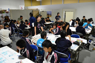 教室内で複数のグループがそれぞれの机で作業をしており、先生たちが巡回して見守っている様子が写っている写真