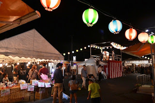 奥に紅白幕が付いたやぐらがあり、屋台のテントやカラフルな提灯が並び、大勢の人々でにぎわう夜の祭り会場の写真