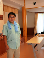 室内で水色のタオルを首にかけて立っている菊地さんの写真