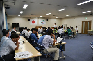 会議室の奥でたくさんの参加者が口の字型に置かれた長机を囲んで座り、手前では前後2列に並んだ長机の席にたくさんの参加者た資料を広げて座っている様子の写真