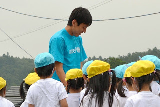 白いシャツを着て黄色や青の帽子をかぶった複数の子供たちが、青いシャツを着た長谷部選手の周りに集まっている写真