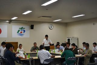 会議室の中で複数の人々が口の字型に置かれたテーブルを囲んで座り、前に立った一人の男性が全員に向かって話をしている様子の写真