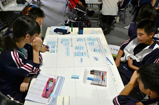 4人の生徒が一つの机を囲み、模造紙の上で資料を見ながら話し合いや書き込みをしている様子が写っている写真