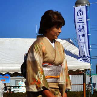 青空の下、南三陸町ののぼり旗が立っている販売ブースの前で、銀色の帯に花柄でベージュ色の着物を着た女性が立っている写真