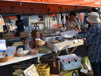 オレンジ色のテントの下の一角で、手編みのかごなどの雑貨商品が棚に陳列されており、女性客が買い物をしている様子の写真