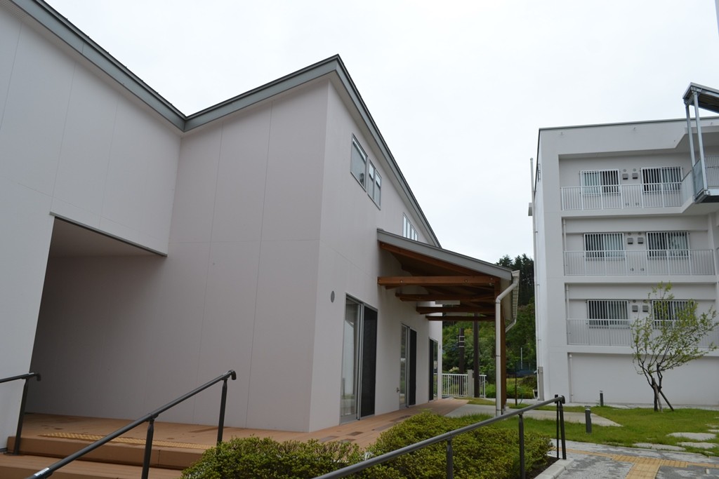 手前に白い壁で木製のデッキと手すり付きの階段がある建物、奥には白い壁で三階建ての建物が建っている写真